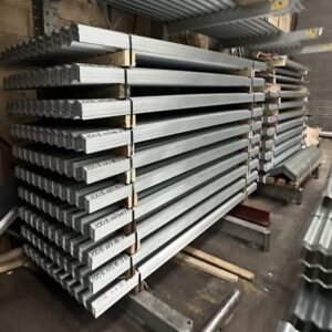 Stacked bundles of galvanised corrugated metal roofing sheets in a warehouse, including flat-packed sheets and wavy-profile roof panels. The silver roofing sheets are strapped on wooden supports and stored for construction or industrial roofing applications.